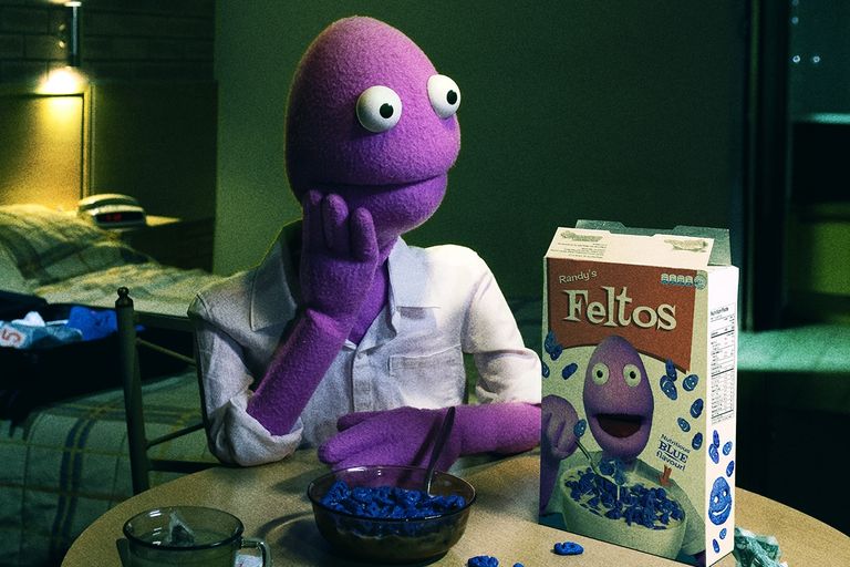 Purple puppet comedian Randy Feltface sitting at a table in a hotel room, resting his head on his hand next to a box of Randy's Feltos cereal.
